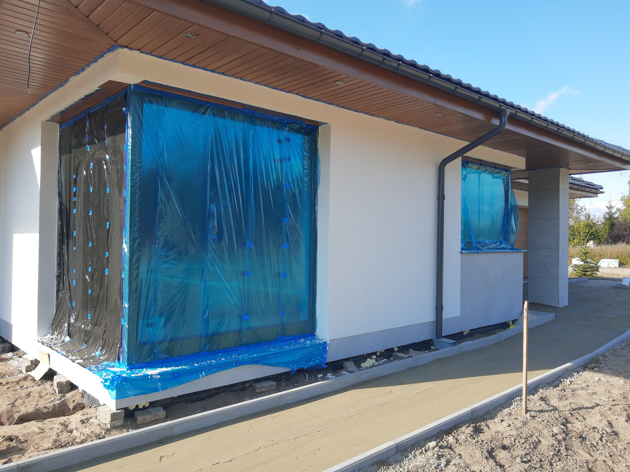 Nowoczesny, parterowy dom w trakcie ocieplania, z oknami zabezpieczonymi folią ochronną, widoczne fundamenty i przygotowane podłoże pod kostkę brukową.