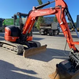 Pomarańczowa minikoparka Kubota na gąsienicach, z łyżką skierowaną do przodu, na betonowym placu, w tle ciężarówka i budynki.