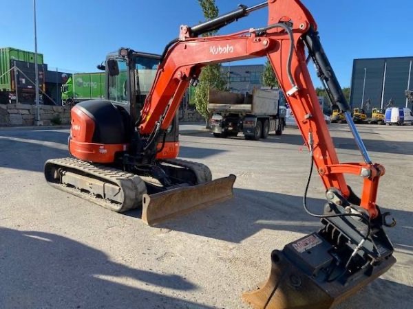 Pomarańczowa minikoparka Kubota na gąsienicach, z łyżką skierowaną do przodu, na betonowym placu, w tle ciężarówka i budynki.