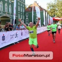 Uśmiechnięty mężczyzna w zielonym stroju biegnie po czerwonym dywanie z podniesionymi rękami podczas maratonu. W tle budynek i kibice.