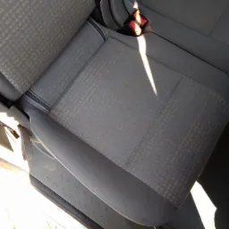 Szary fotel samochodowy z delikatnym wzorem w kratkę, oświetlony jasnym światłem słonecznym wpadającym przez otwarte drzwi.