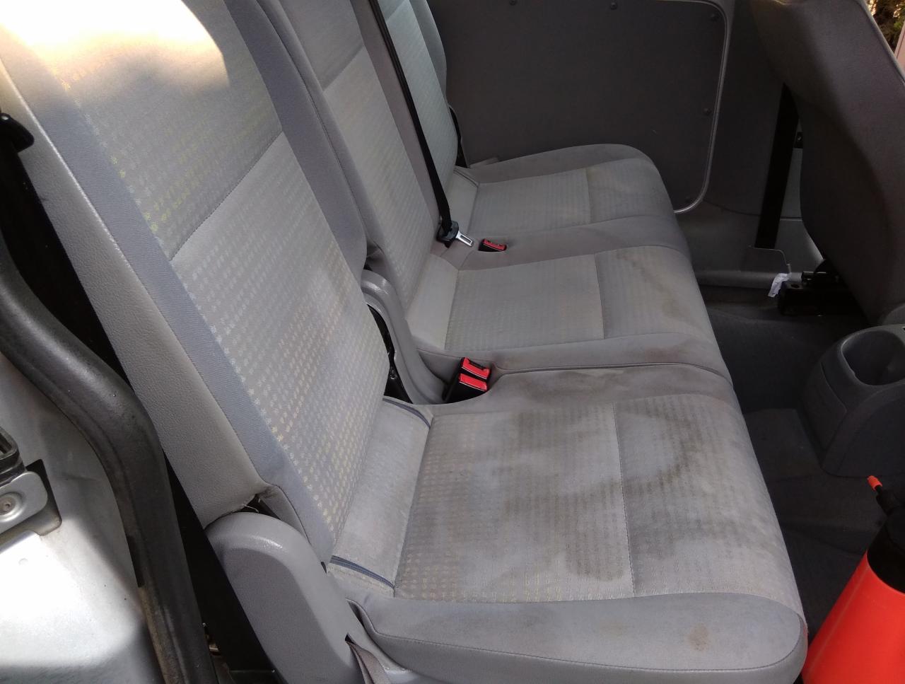 Wnętrze samochodu z widocznymi, szarymi fotelami z plamami, prawdopodobnie po czyszczeniu, z widocznym czerwonym spryskiwaczem w prawym dolnym rogu.