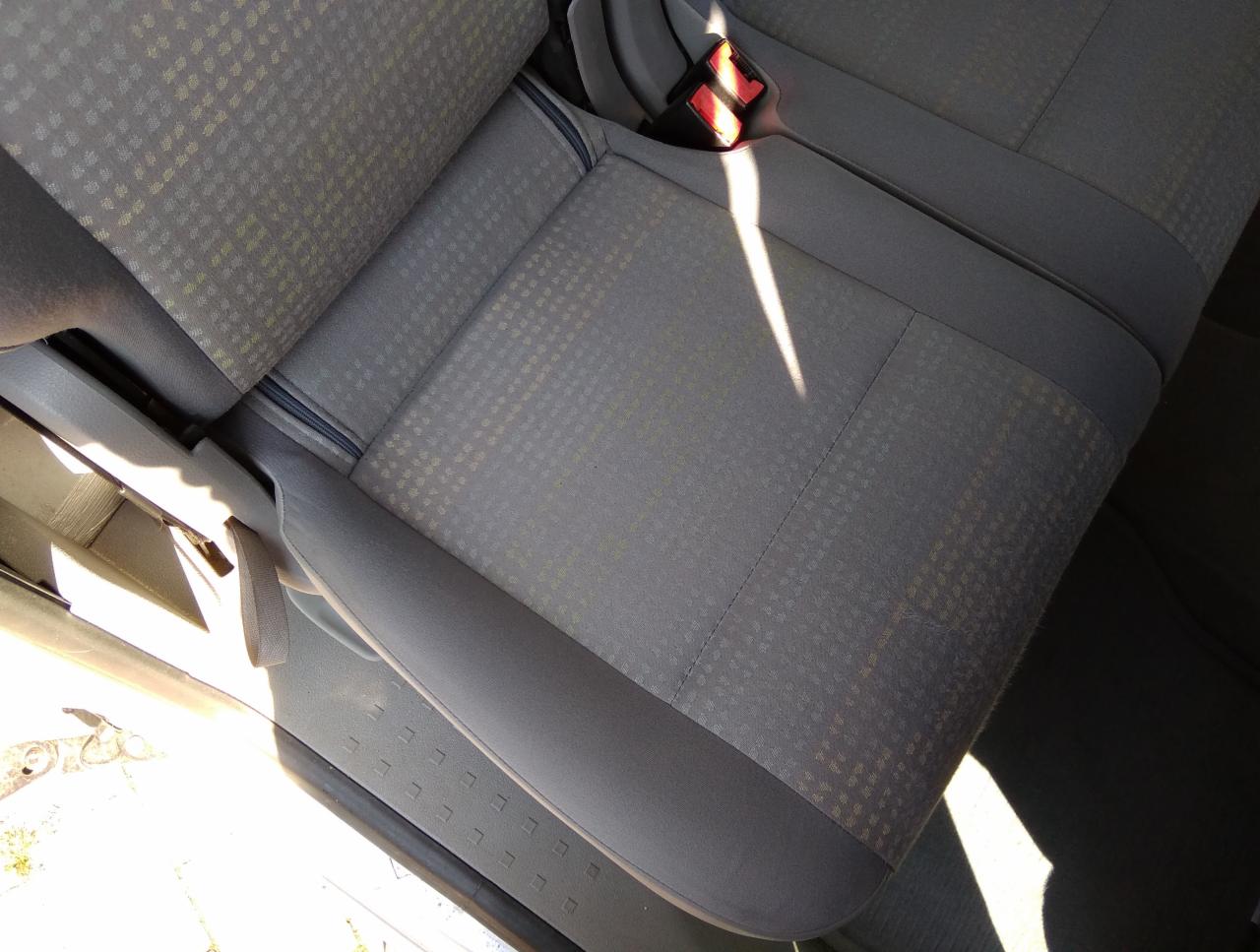 Szary fotel samochodowy z delikatnym wzorem w kratkę, oświetlony jasnym światłem słonecznym wpadającym przez otwarte drzwi.