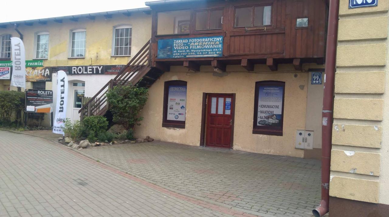 Fasada budynku z szyldami reklamowymi i informacyjnymi różnych firm, w tym zakładu fotograficznego 'Arenika' i pośrednictwa ubezpieczeniowego, widoczne także reklamy rolet i żaluzji.