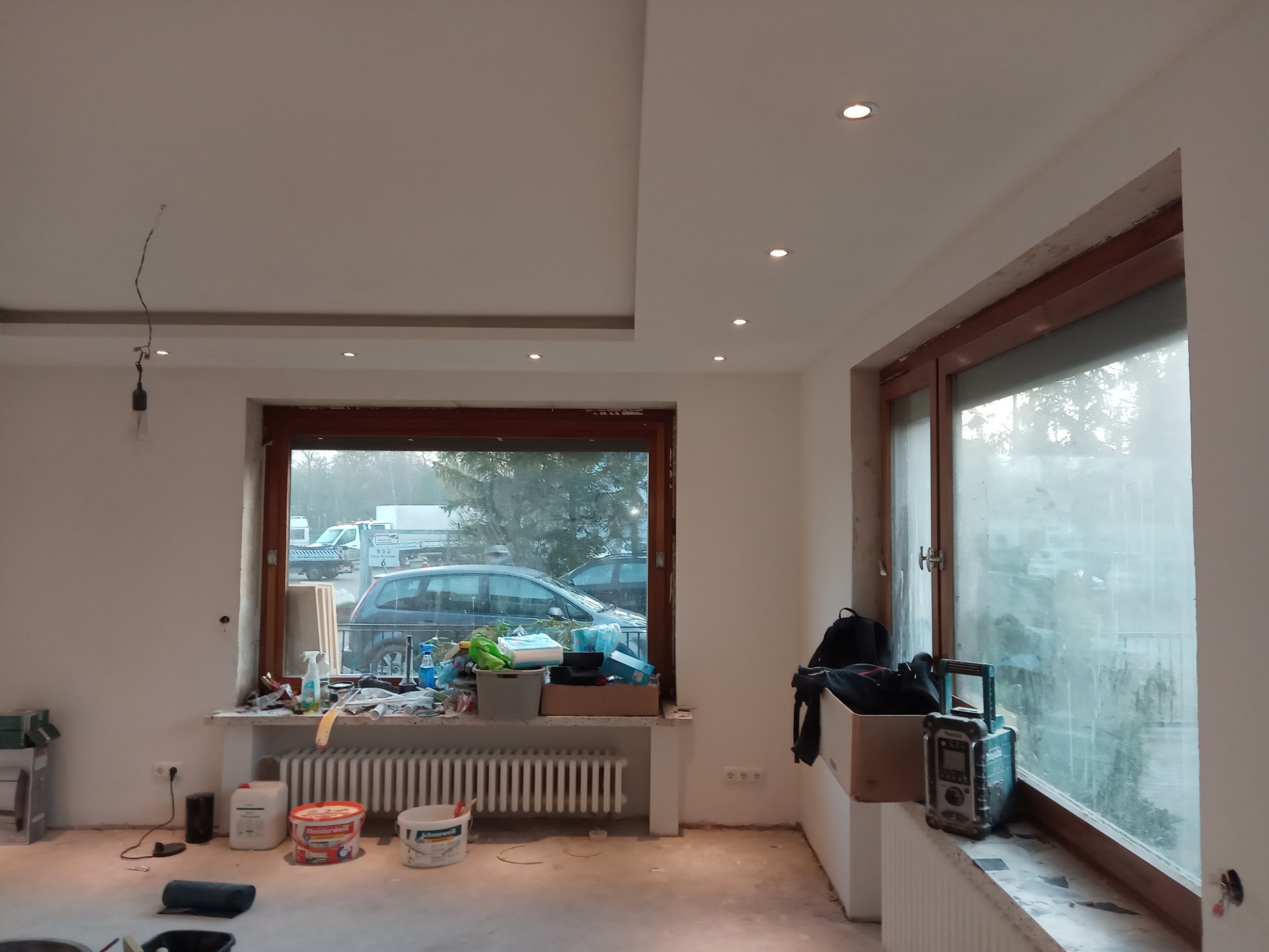 Pokój w trakcie remontu: świeżo pomalowane ściany, sufit z oświetleniem punktowym, okna z widokiem na zewnątrz, farby i narzędzia malarskie zgromadzone pod oknem.