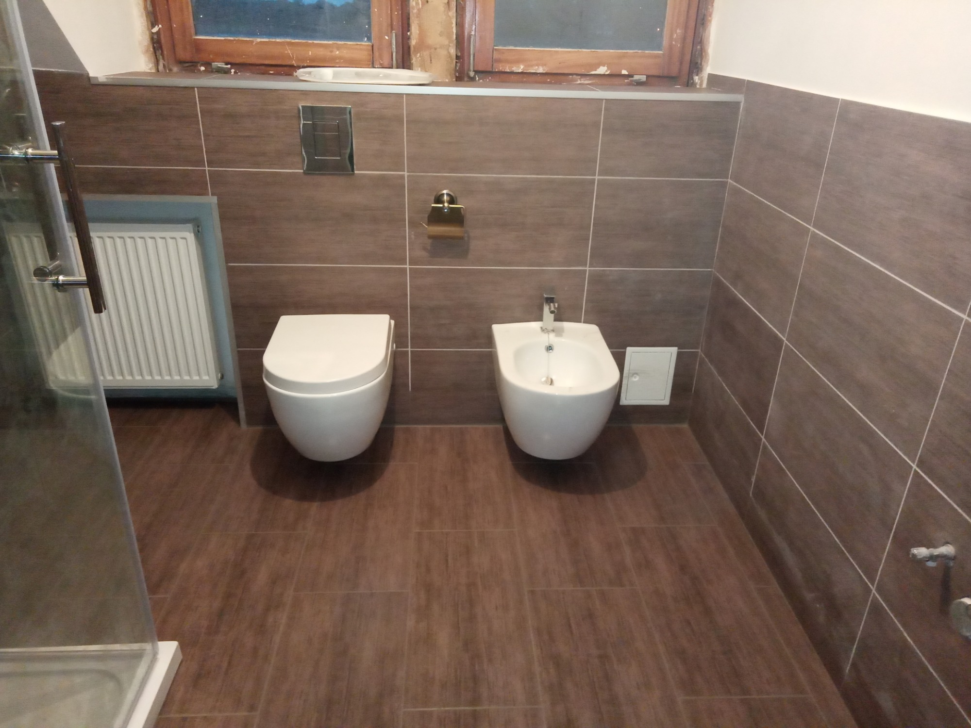 Nowoczesna łazienka z brązowymi płytkami imitującymi drewno, wisząca toaleta i bidet, chromowane elementy armatury, częściowo widoczna kabina prysznicowa.