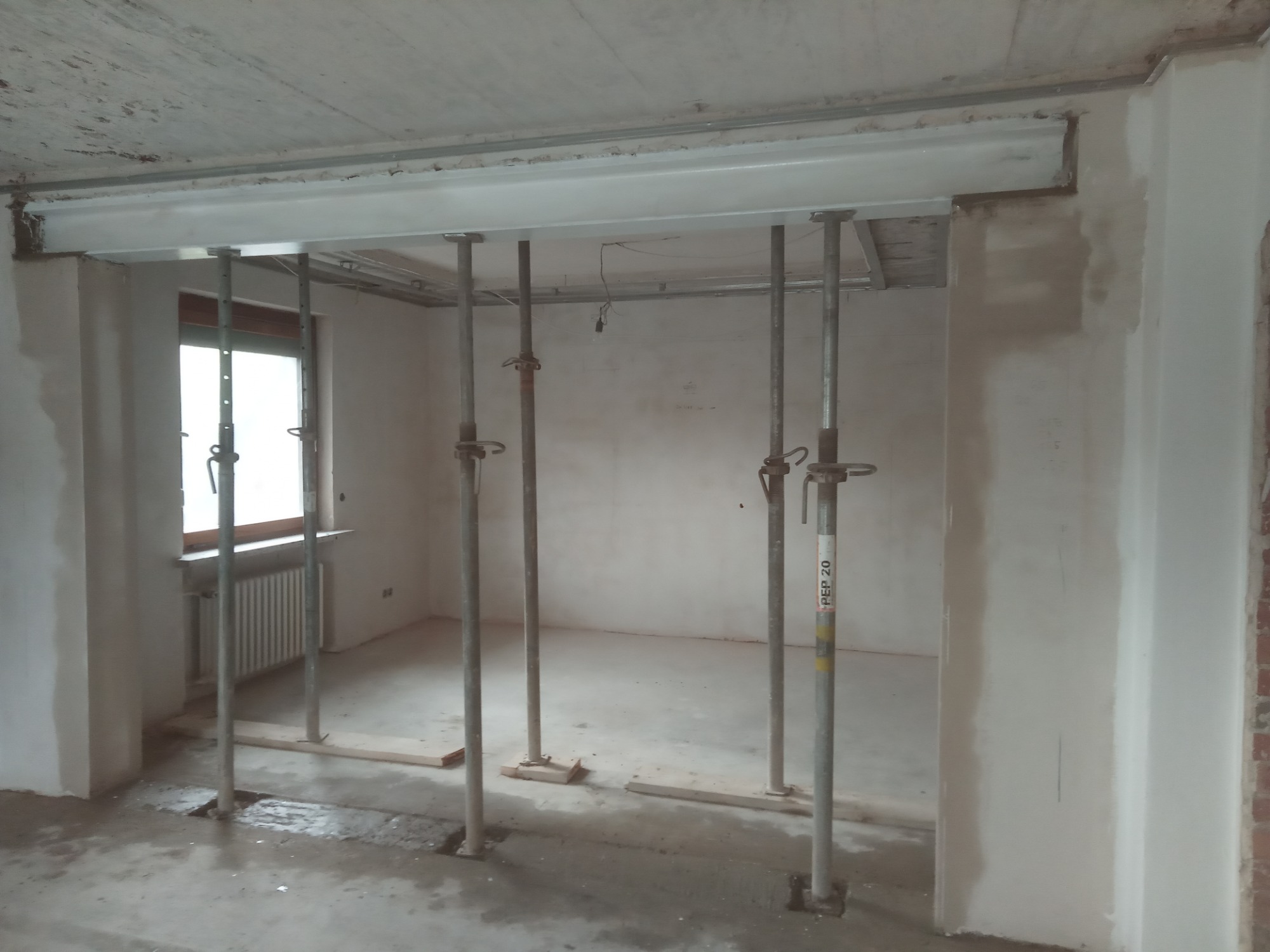 Wnętrze w trakcie prac z widocznymi stalowymi podporami stropowymi, drewnianymi podkładkami i metalowym profilem pod sufitem, ściany w surowym stanie.