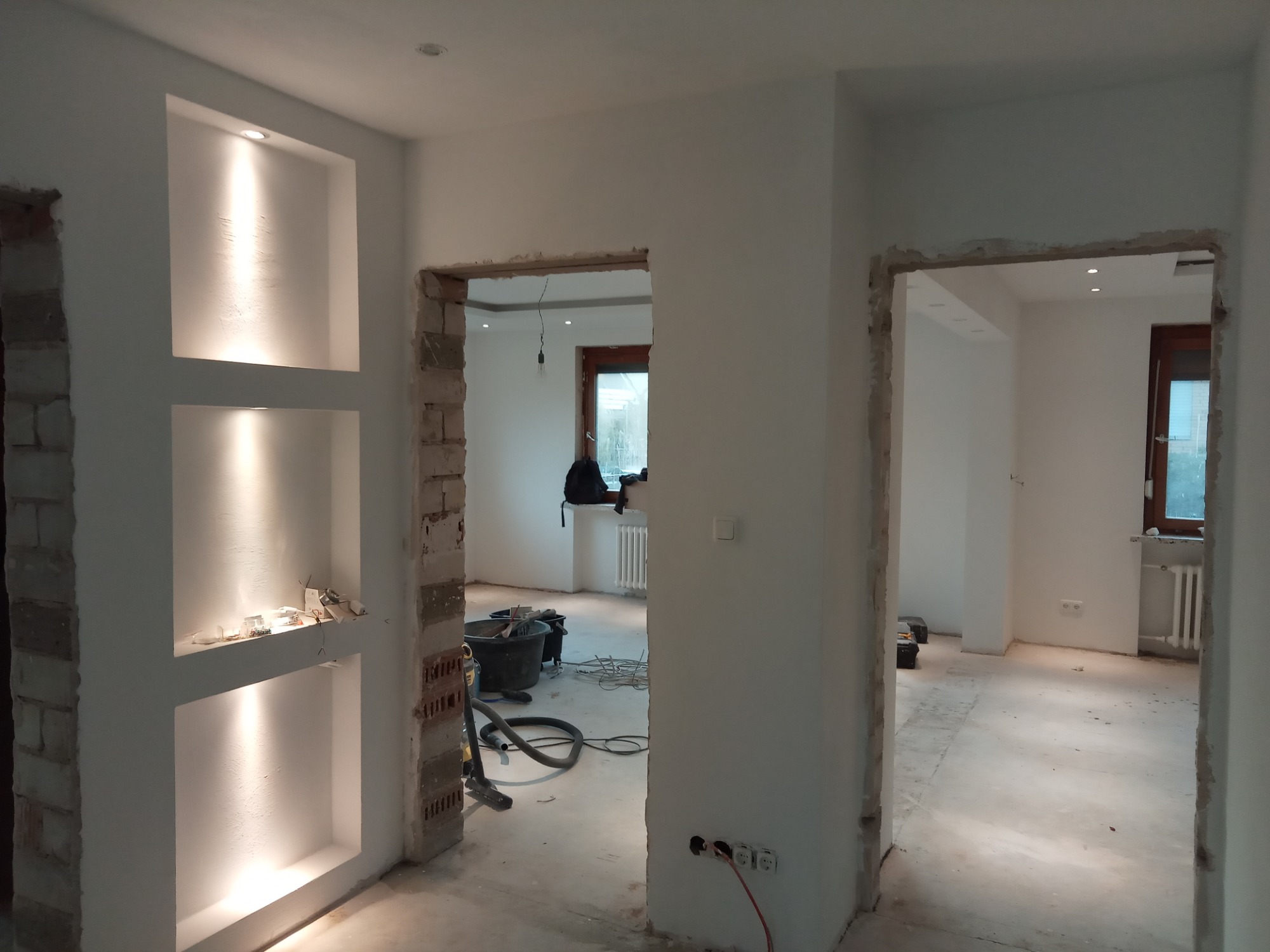 Wnętrze w trakcie prac wykończeniowych: odkryta cegła przy wnękach oświetleniowych, widoczne kable, gniazdka i narzędzia budowlane, ściany przygotowane do malowania.
