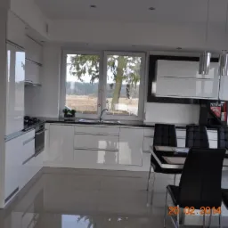 Nowoczesna, biała kuchnia z połyskiem, z czarnym blatem i widokiem na zewnątrz przez duże okno. W tle widoczny automat do kawy Nescafé.