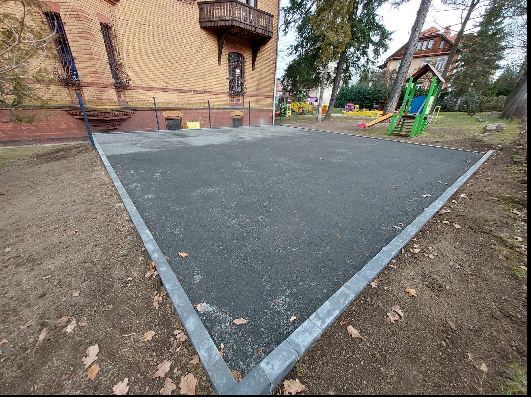 Nowa, czarna, gumowa nawierzchnia placu zabaw w Jeżowie Sudeckim, obrzeża wykończone szarą kostką, w tle budynek z cegły i elementy placu zabaw.