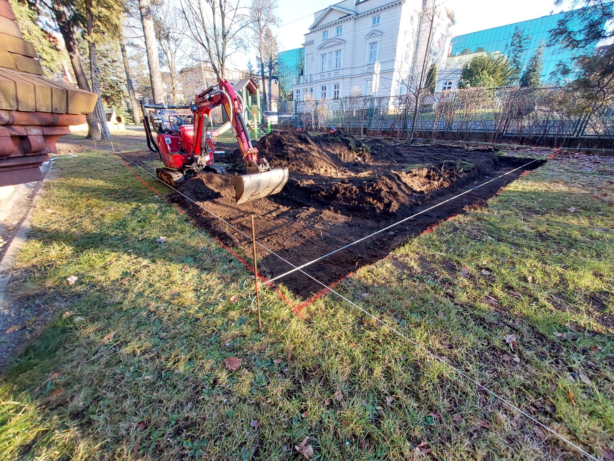 Czerwony minikoparka Yanmar przygotowuje teren pod trawnik w ogrodzie, wykopana ziemia obok, wyznaczony obszar sznurkiem, w tle budynek.