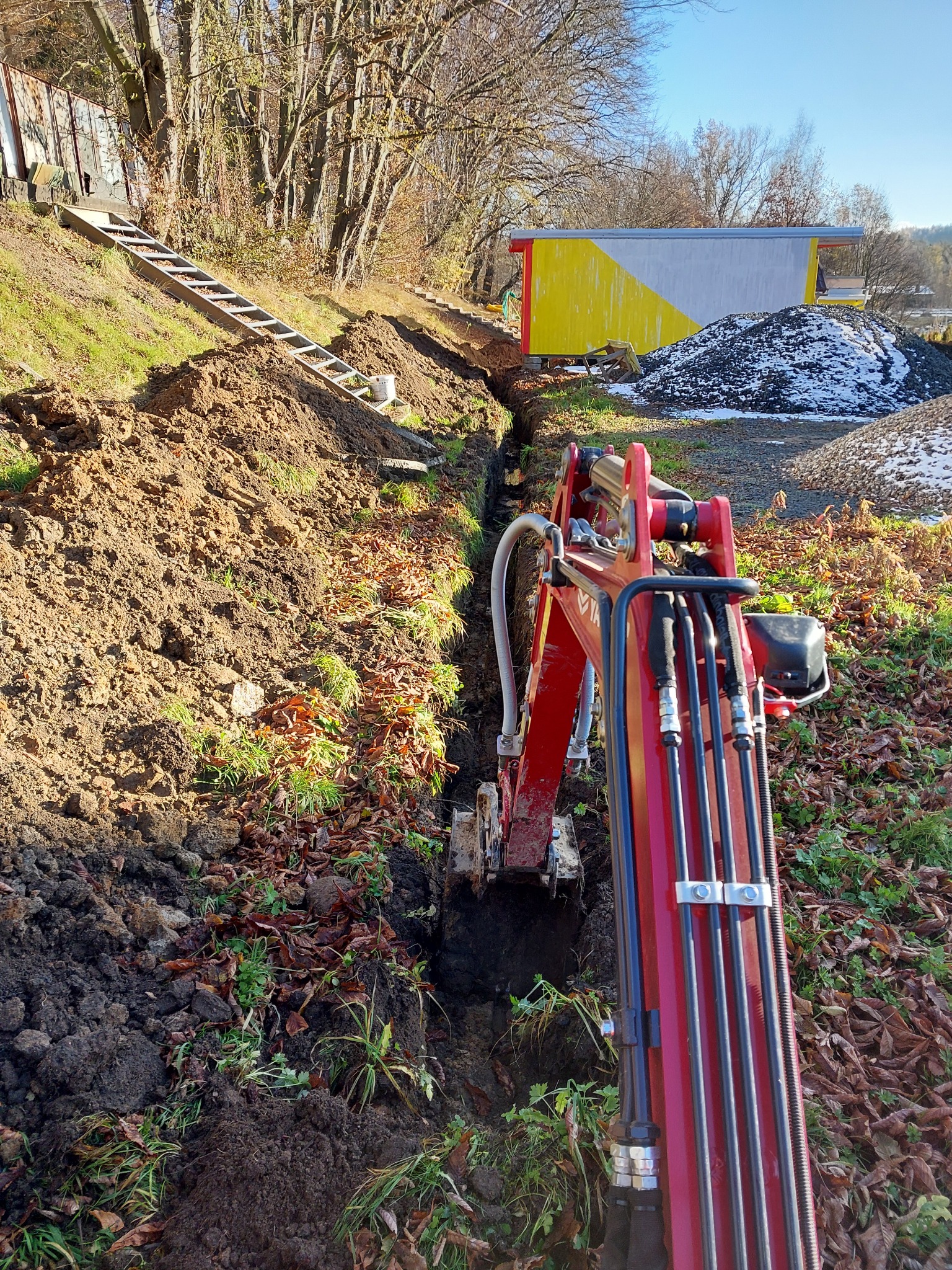Czerwona minikoparka wykopująca wąski rów w ziemi na działce z drzewami, w tle widoczny budynek z żółto-białą ścianą i sterta gruzu.