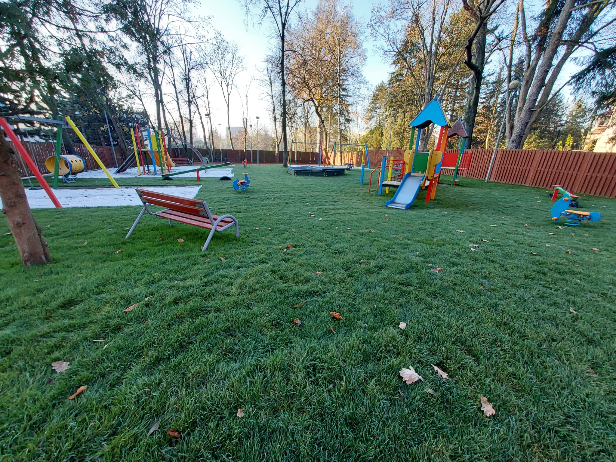 Widok na trawnik z placem zabaw w tle, z ławką na pierwszym planie i opadłymi liśćmi na trawie.