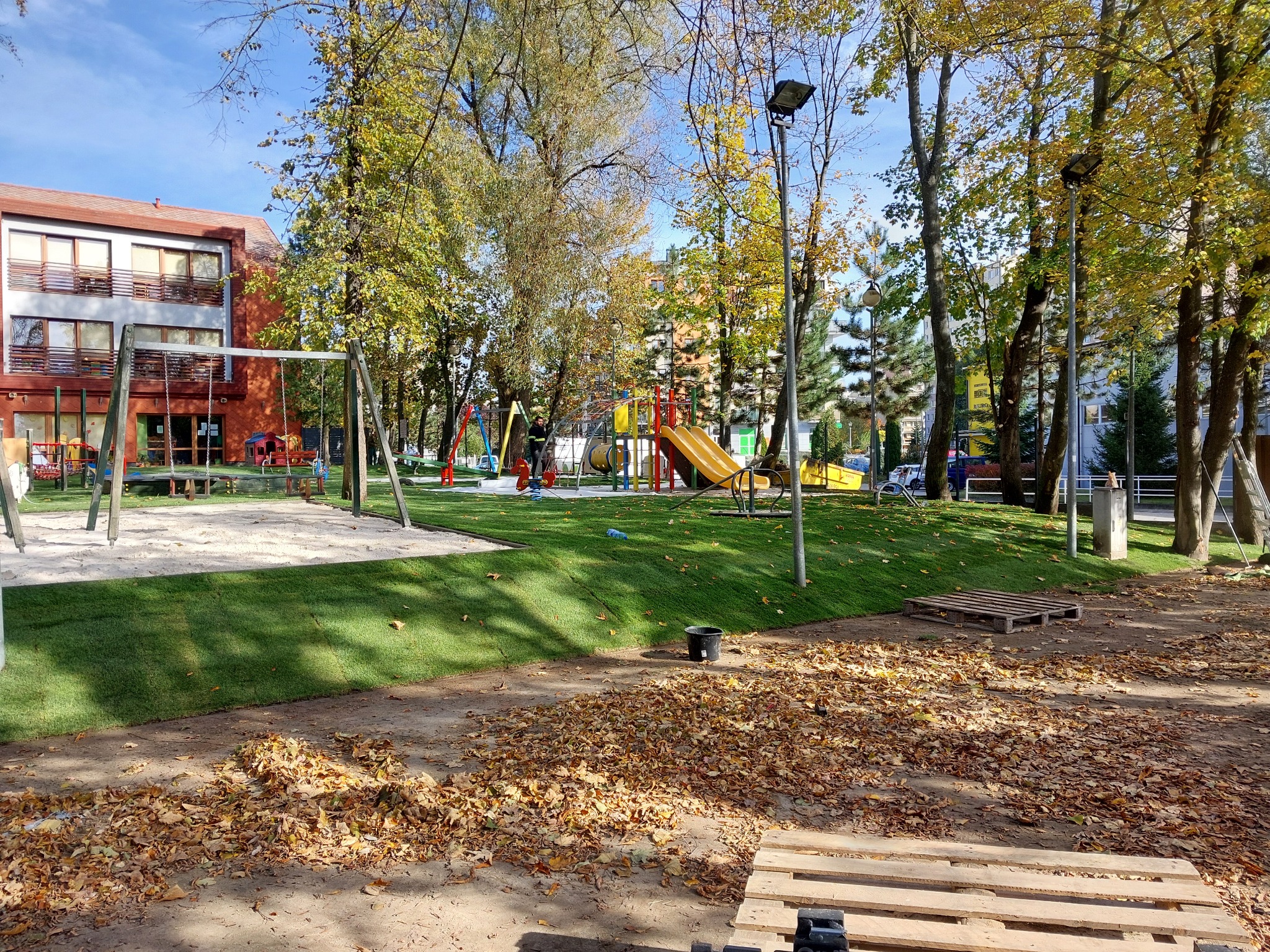 Nowo założony trawnik z rolki na placu zabaw w jesiennej scenerii, z widocznymi paletami i opadłymi liśćmi.