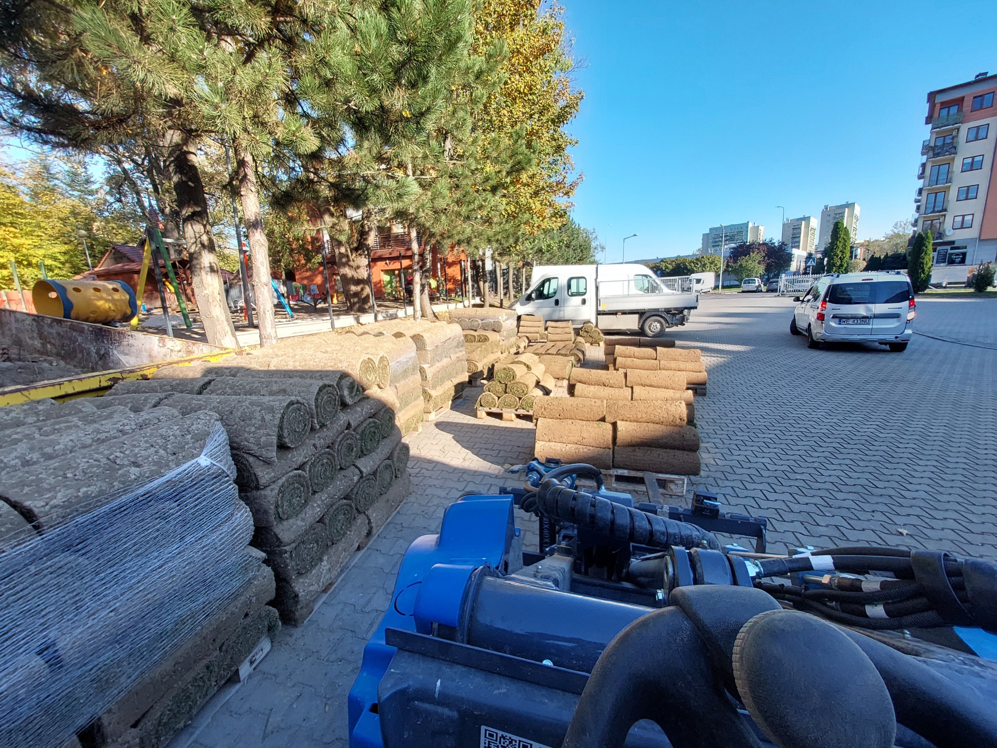 Stosy trawy w rolkach na paletach, przygotowane do rozłożenia, widoczne z perspektywy maszyny do rozkładania, na tle placu zabaw i budynków mieszkalnych w słoneczny dzień.