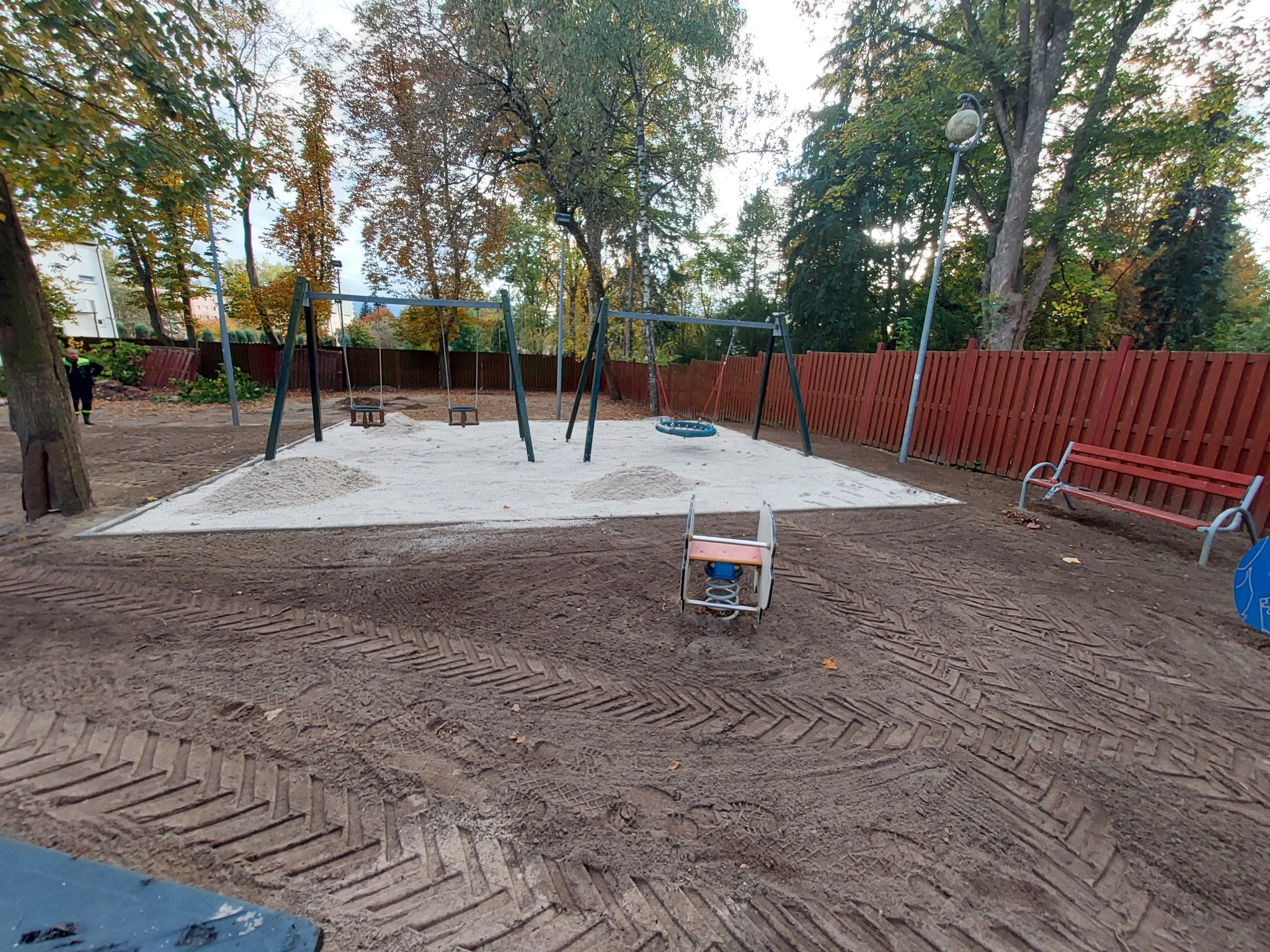 Nowo wybudowany plac zabaw z huśtawkami, piaskiem, czerwoną ławką i ogrodzeniem na obrzeżach, widoczne ślady opon po maszynach budowlanych.