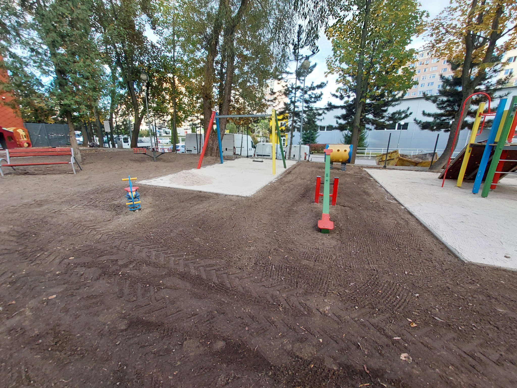 Świeżo przygotowany teren pod plac zabaw z widocznymi śladami opon i piaskiem, otoczony drzewami i budynkami w tle.