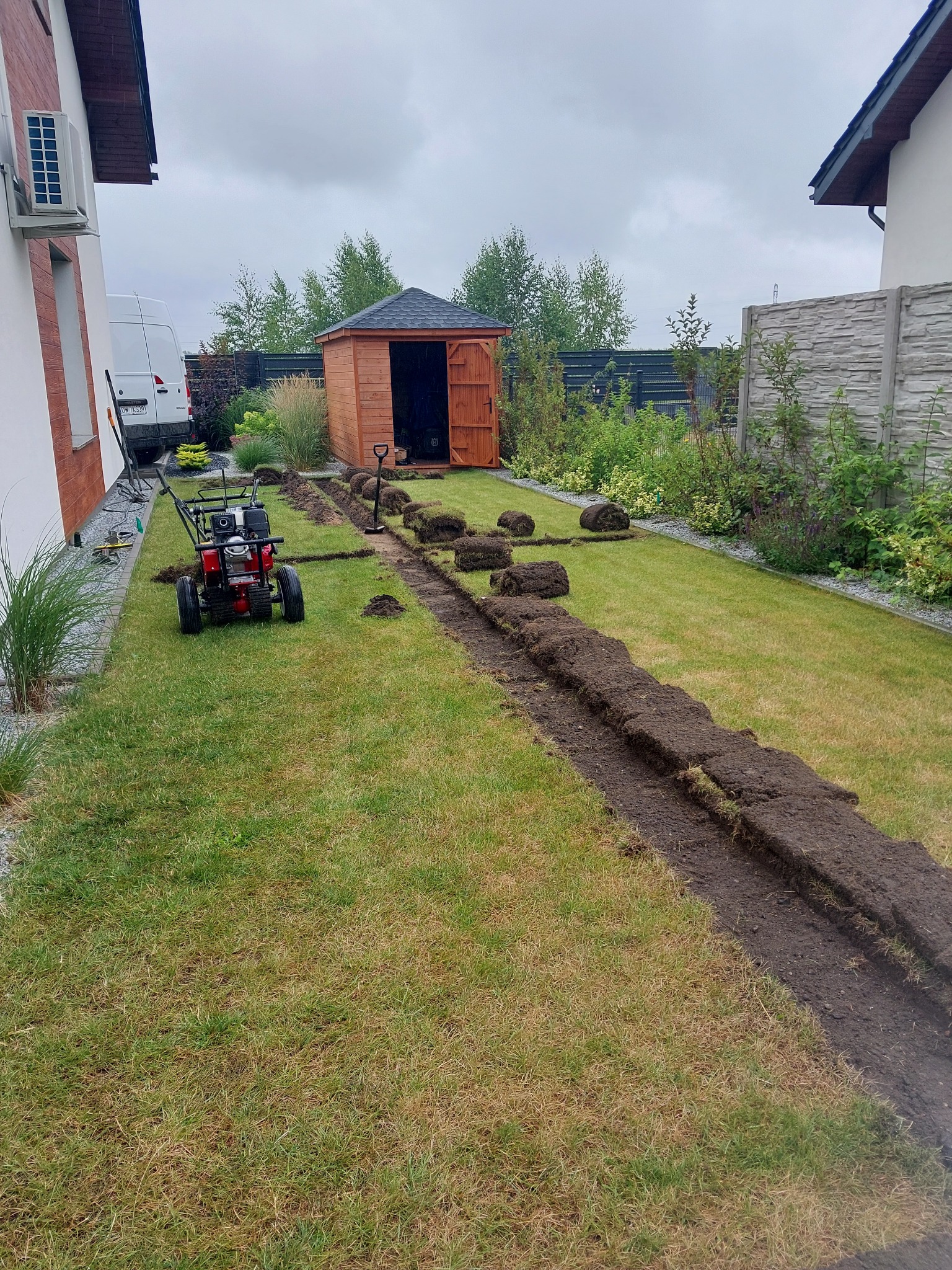 Przygotowanie terenu pod trawnik z rolki: wykopany rów, obok zwinięte rolki trawy, glebogryzarka i szpadel w tle, widok na ogród z domkami.