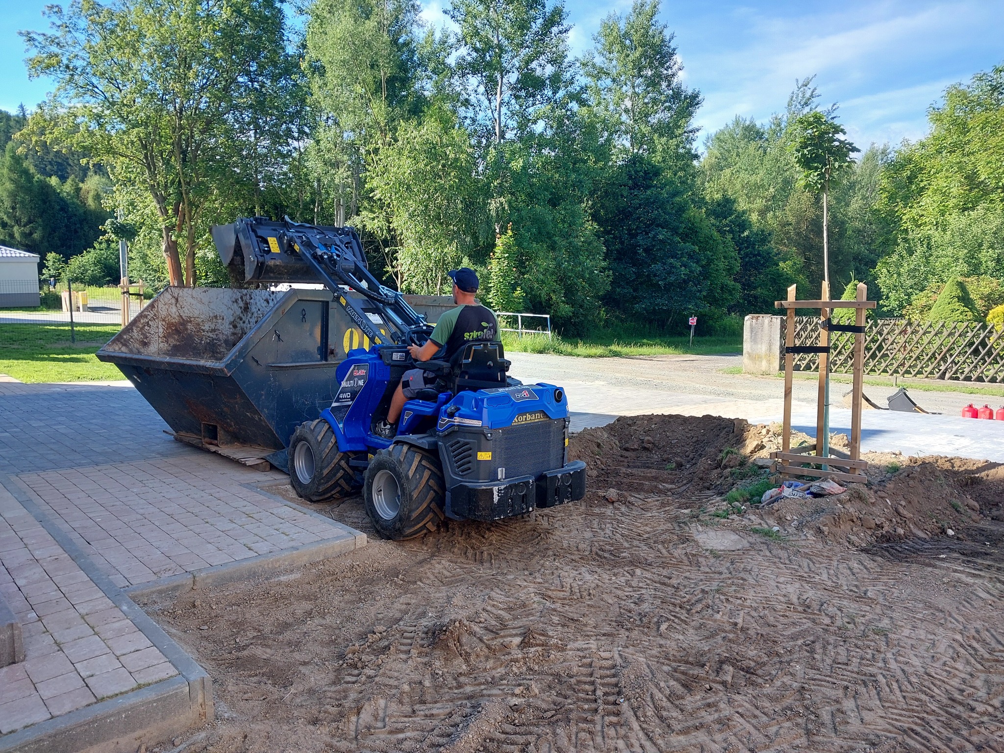 Pracownik na niebieskiej miniładowarce z łyżką, transportujący metalowy kontener z ziemią na tle zieleni i kostki brukowej, świeżo posadzone drzewko z drewnianą podporą obok.