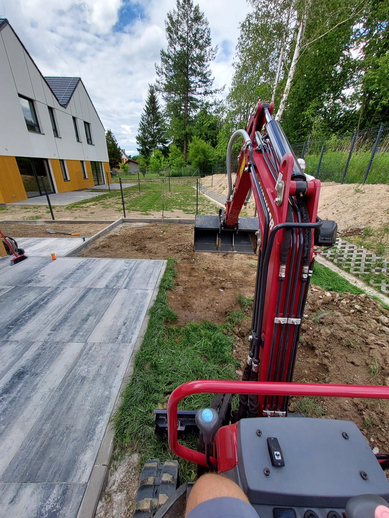 Czerwona mini koparka Yanmar przygotowuje teren pod trawnik z rolki obok nowo ułożonego chodnika z szarych płyt betonowych przy nowoczesnym domu z żółtymi akcentami.