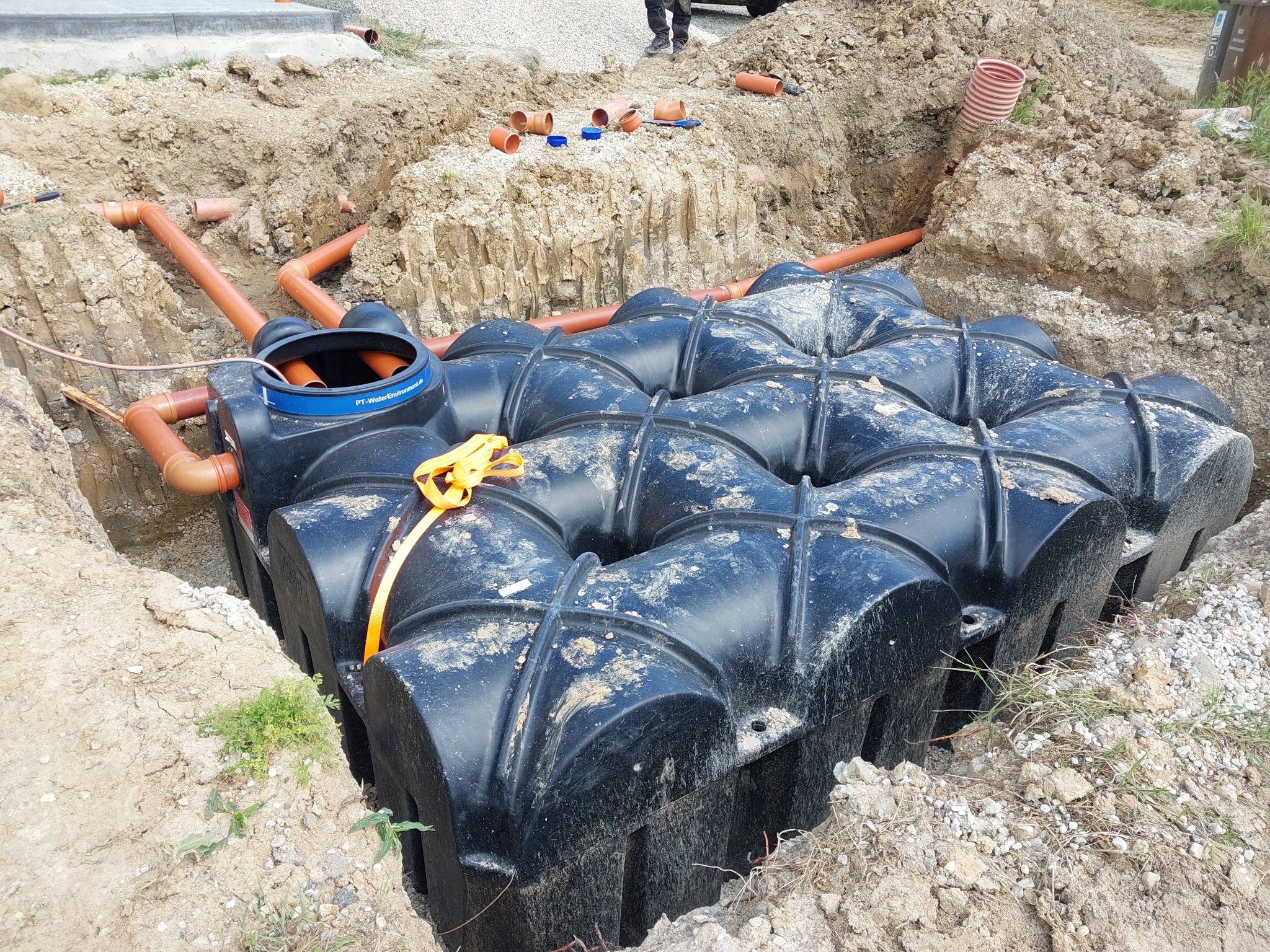 Czarny zbiornik retencyjny modułowy zakopany w wykopie, z podłączonymi pomarańczowymi rurami kanalizacyjnymi, zabezpieczony żółtą taśmą, widoczne narzędzia i dodatkowe rury na górze wykopu.