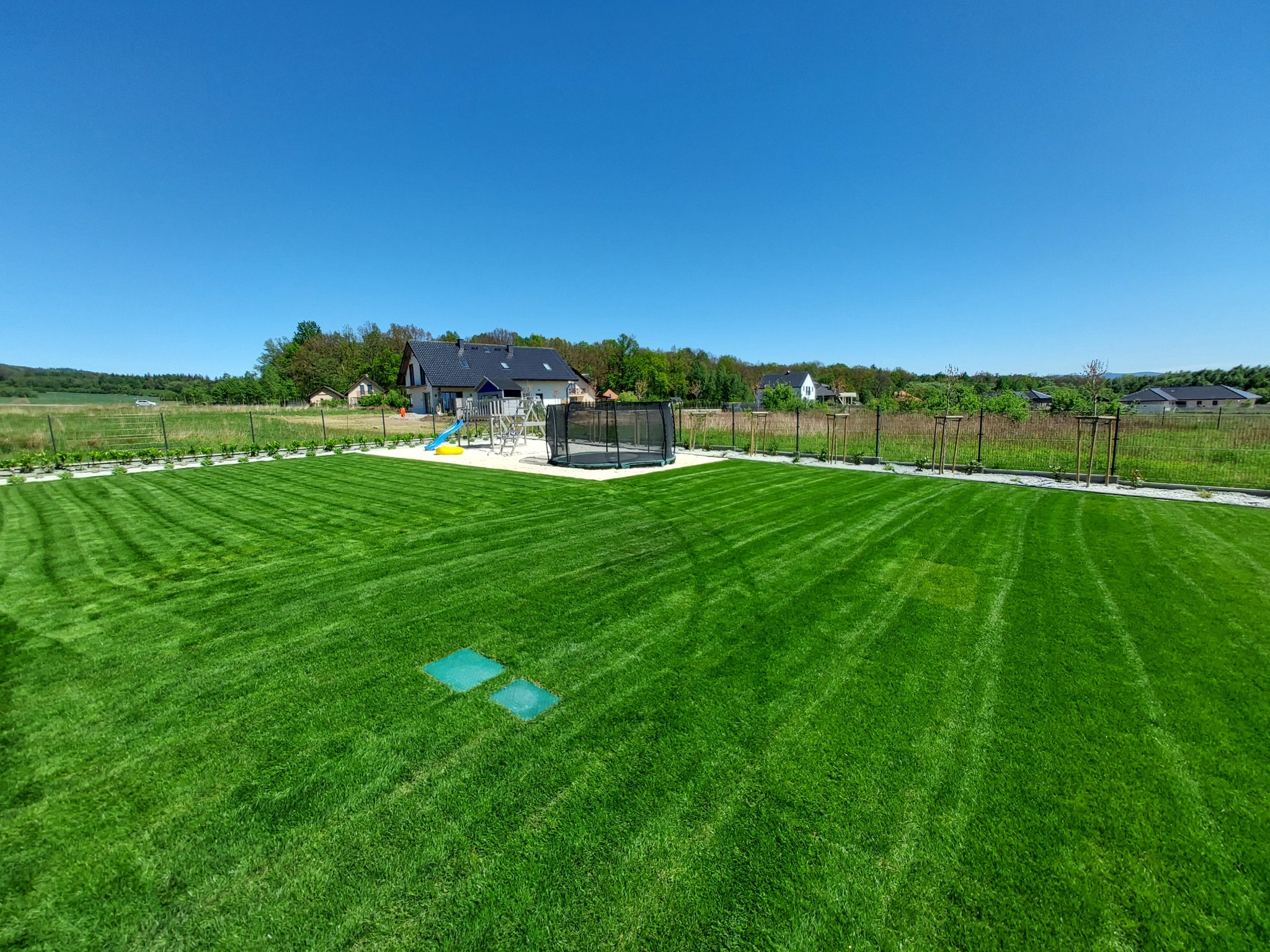 Idealnie przystrzyżony trawnik z widocznymi pasami po kosiarce, z dwoma zielonymi włazami w trawie, trampoliną i placem zabaw w tle, otoczony niskim ogrodzeniem.