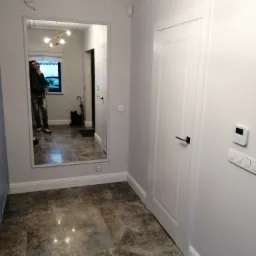 Wnętrze przedpokoju po odświeżeniu: szare ściany, białe drzwi z czarną klamką, duże lustro z odbiciem osoby w ubraniach roboczych i nowoczesna lampa sufitowa.