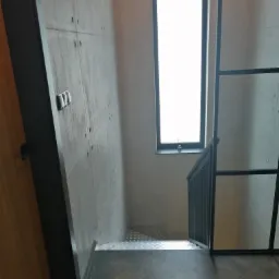 Wnętrze klatki schodowej z betonową ścianą imitującą szalunek, parkietem ułożonym w jodełkę, metalowymi schodami i czarną stolarką okienną.