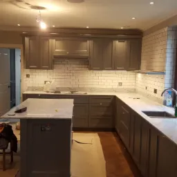 Szare szafki kuchenne z białym marmurowym blatem i białymi płytkami metra, widoczne ślady prac wykończeniowych, podłoga zabezpieczona kartonem.