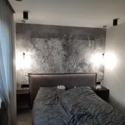 Sypialnia z łóżkiem, szafkami nocnymi i dekoracyjną ścianą z motywem roślinnym, oświetlona lampami wiszącymi i sufitowymi.