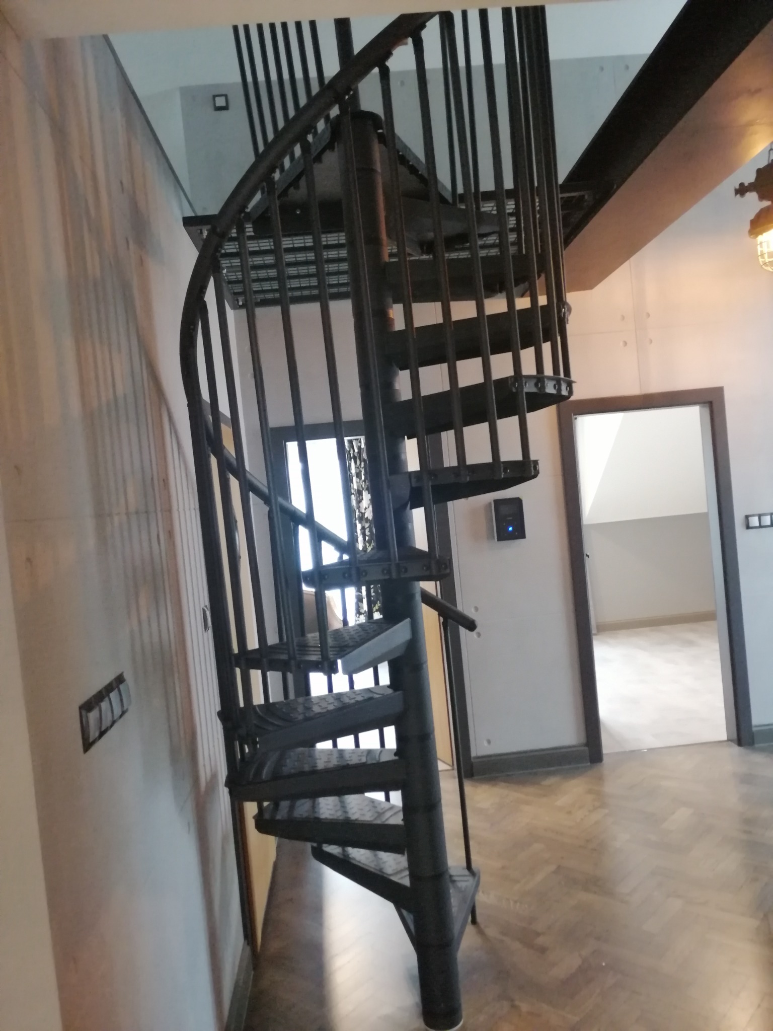 Czarna, metalowa klatka schodowa spiralna w nowoczesnym wnętrzu, widok na drewnianą podłogę ułożoną w jodełkę i drzwi do innego pomieszczenia.