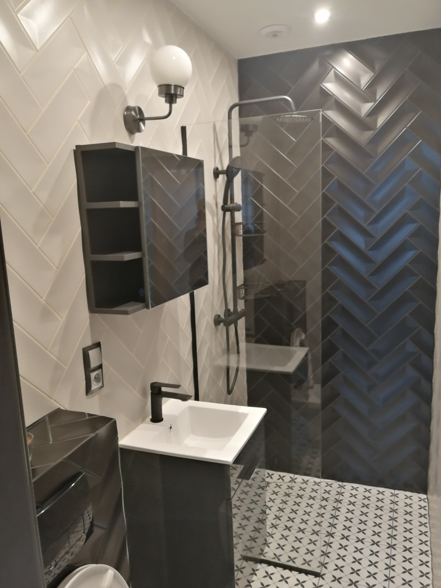 Nowoczesna łazienka z szarymi płytkami ułożonymi w jodełkę na ścianach i pod prysznicem, białą umywalką z czarną armaturą, szafką z lustrem i wzorzystą podłogą.