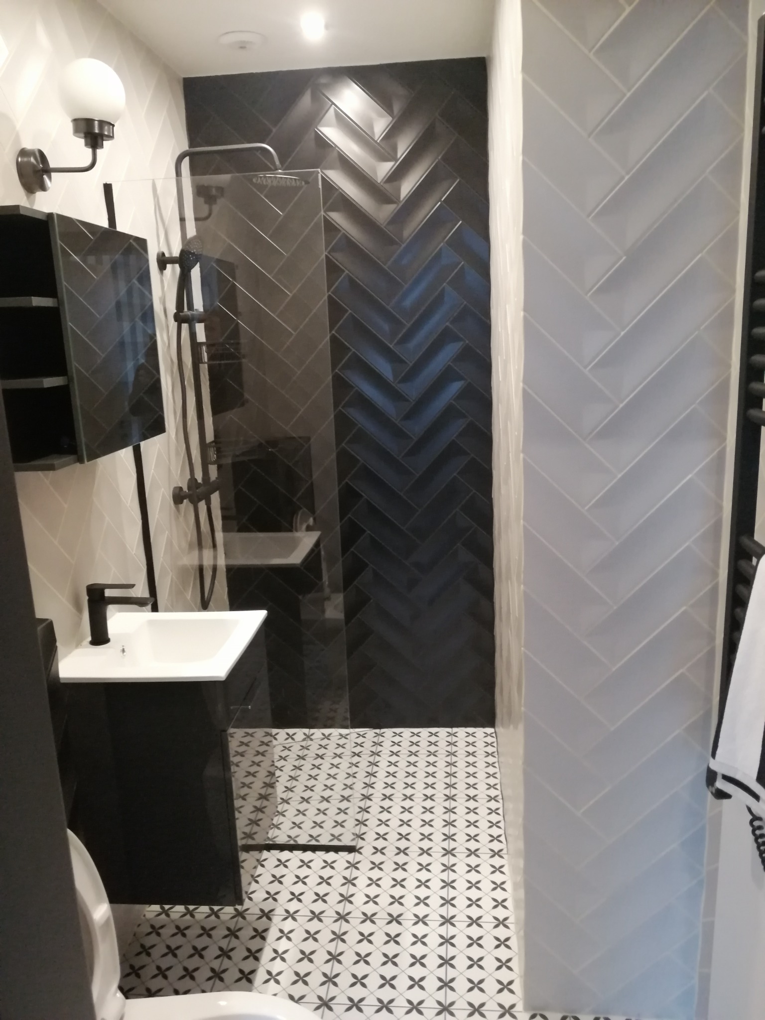 Nowoczesna łazienka z czarnymi i białymi płytkami ułożonymi w jodełkę na ścianach i podłodze, czarna armatura i szafka pod umywalką, szklana kabina prysznicowa.