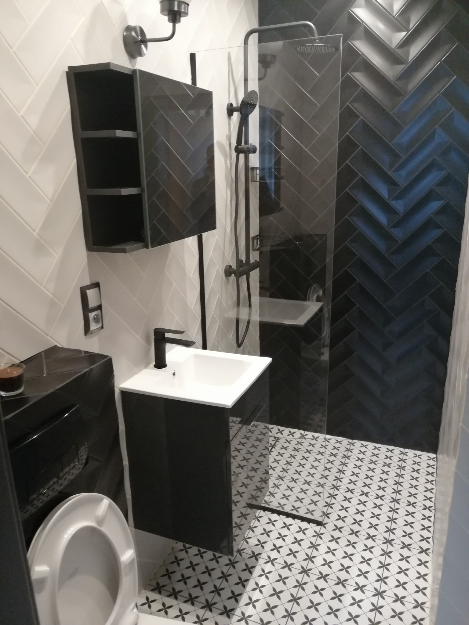 Nowoczesna łazienka z czarno-białymi płytkami ułożonymi w jodełkę na ścianie prysznica i wzorzystą podłogą, minimalistyczna umywalka z czarną armaturą i szafka wisząca z otwartymi półkami.