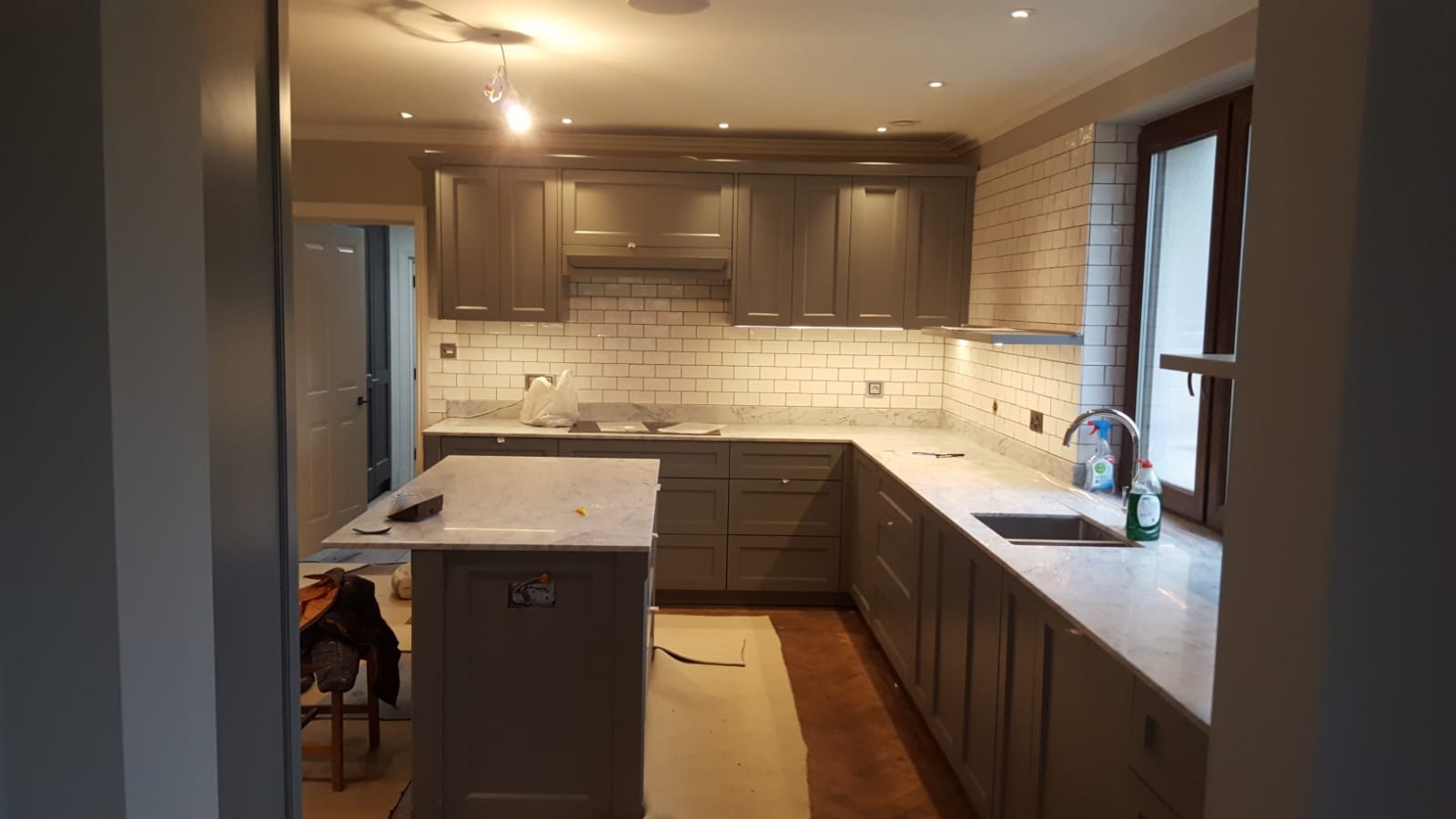 Szare szafki kuchenne z białym marmurowym blatem i białymi płytkami metra, widoczne ślady prac wykończeniowych, podłoga zabezpieczona kartonem.