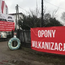 Czerwony baner z napisem 'OPONY WULKANIZACJA' zawieszony na ogrodzeniu warsztatu samochodowego, obok zużyta opona pomalowana na niebiesko-zielono z napisem 'WULKANIZACJA', w tle budynek warsztatu...