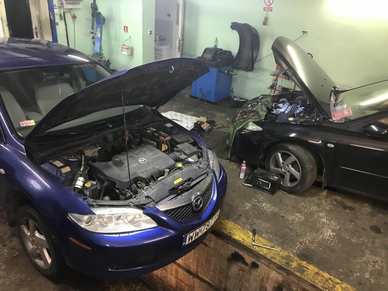 Dwa samochody Mazda z otwartymi maskami w warsztacie; niebieski z przodu, czarny z tyłu, widoczne silniki i narzędzia; podłoga warsztatu z widocznymi śladami użytkowania, ściany w odcieniach zieleni.