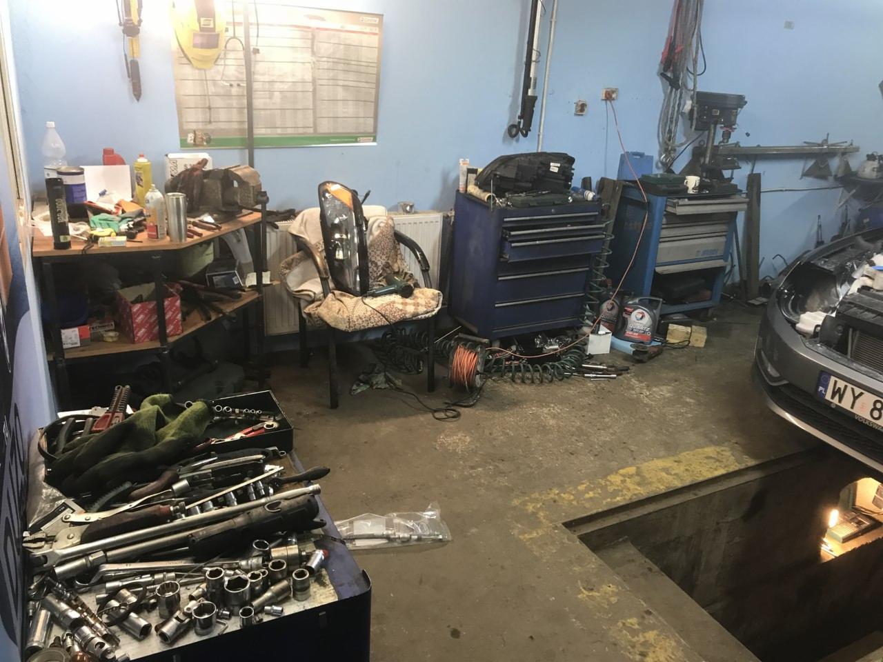 Wnętrze warsztatu samochodowego z widocznym kanałem, narzędziami na stole, szafką narzędziową i częściowo rozebranym samochodem z otwartą maską, na krześle leży reflektor samochodowy.