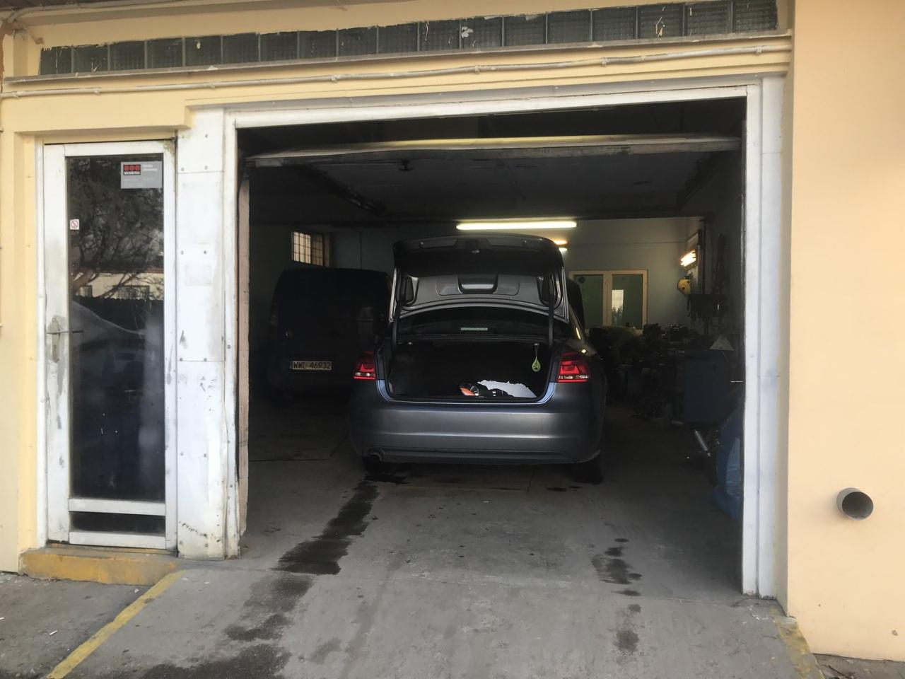 Wnętrze warsztatu samochodowego z otwartym bagażnikiem szarego sedana, widoczne narzędzia i inne pojazdy w tle.