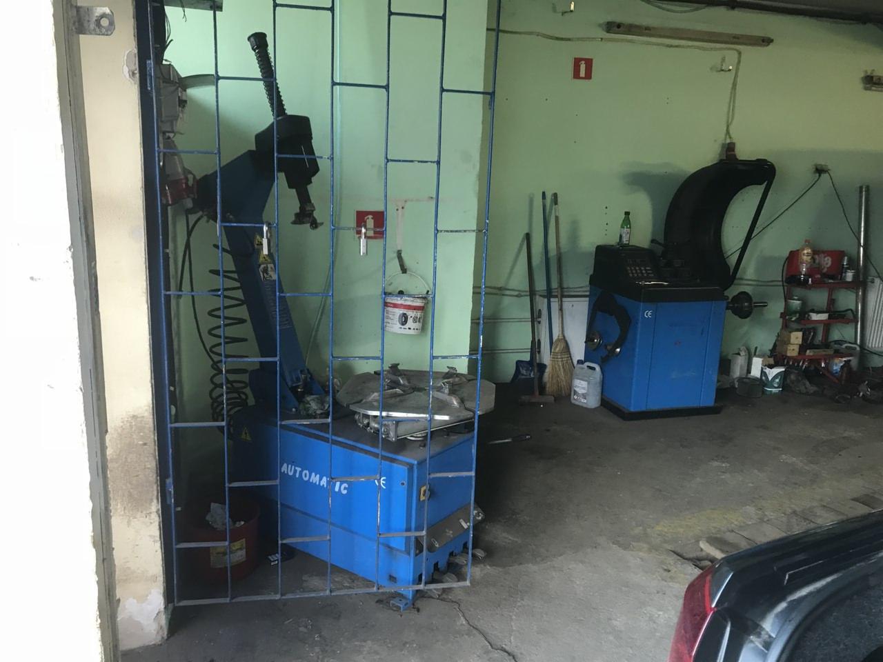 Wnętrze warsztatu samochodowego z maszyną do zmiany opon za niebieską kratą i wyważarką kół w tle, widoczne narzędzia i akcesoria.