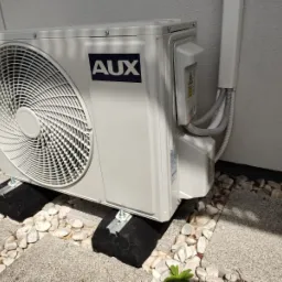 Zewnętrzna jednostka klimatyzacji marki AUX umieszczona na czarnych, amortyzujących podkładkach na tle białego muru i kamiennego podłoża z białymi otoczakami.