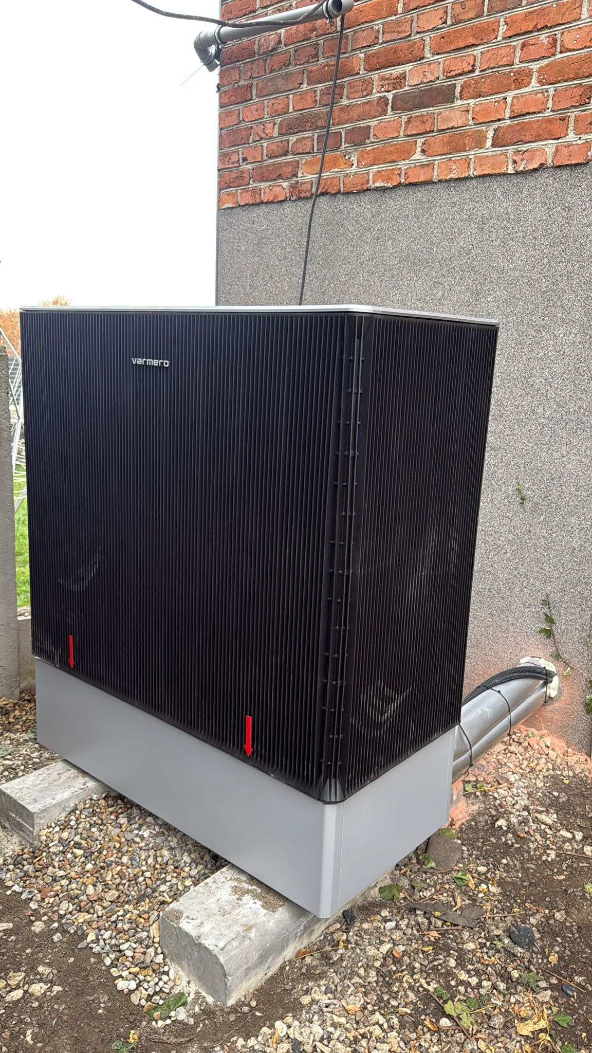 Czarna pompa ciepła Varmero na betonowych blokach, z widocznymi rurami i fragmentem ceglanego budynku w tle. Ujęcie z dołu, podkreślające kubaturę urządzenia.