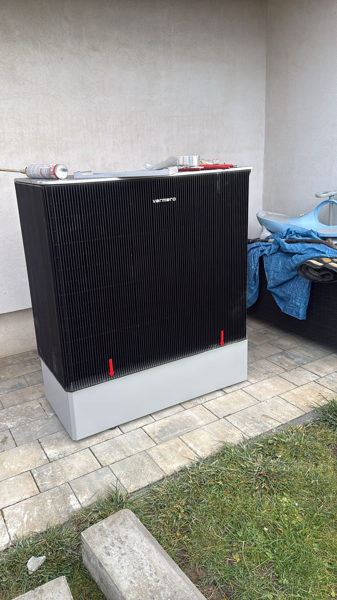 Zewnętrzna pompa ciepła marki Varmero, ustawiona na kostce brukowej przy ścianie budynku. Widoczne narzędzia montażowe na górze urządzenia. Trawa w tle.