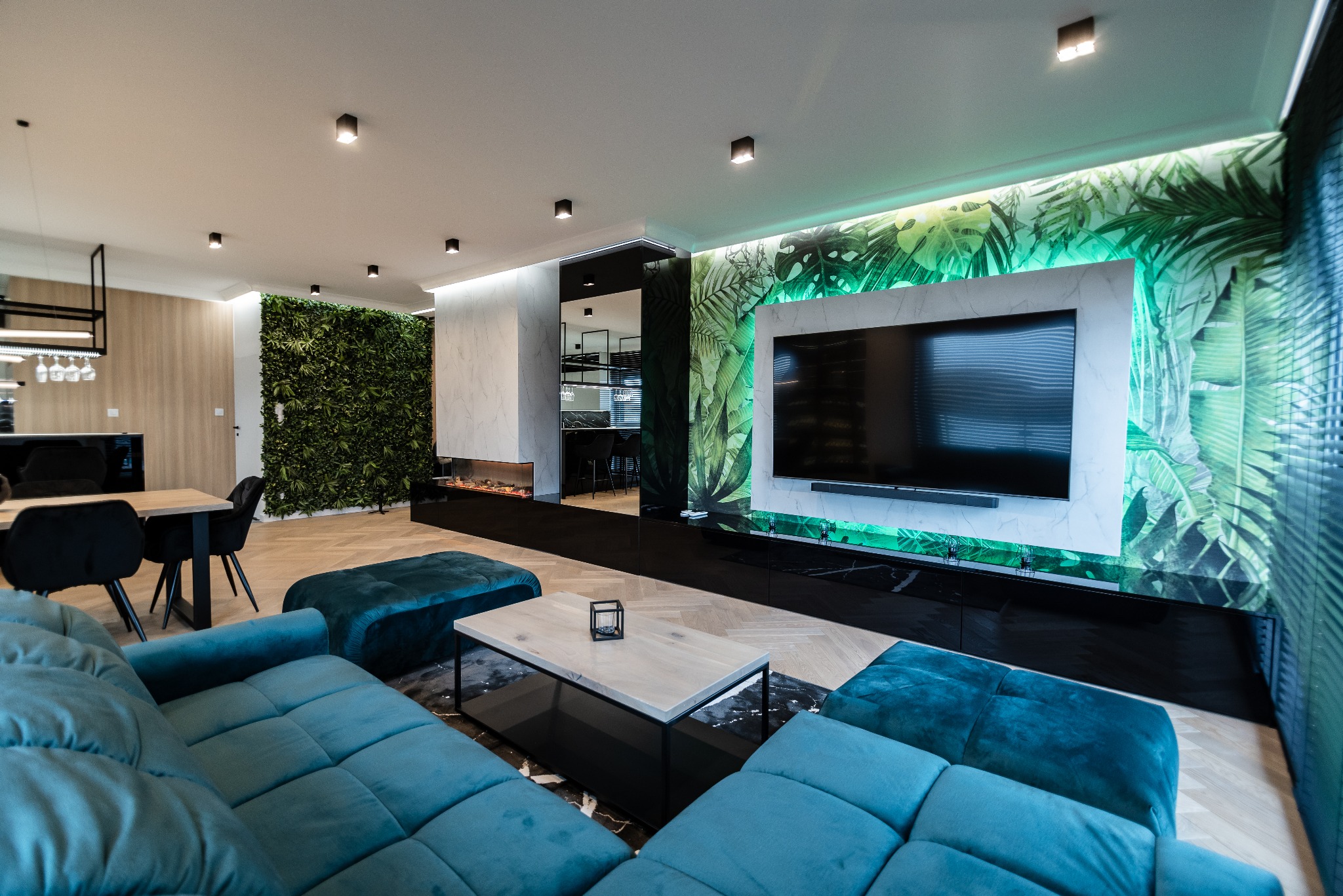 Salon z zieloną ścianą i tapetą w liście, turkusowa sofa, drewniana podłoga. Nowoczesne oświetlenie podkreśla tropikalny motyw. Widok na jadalnię w tle.