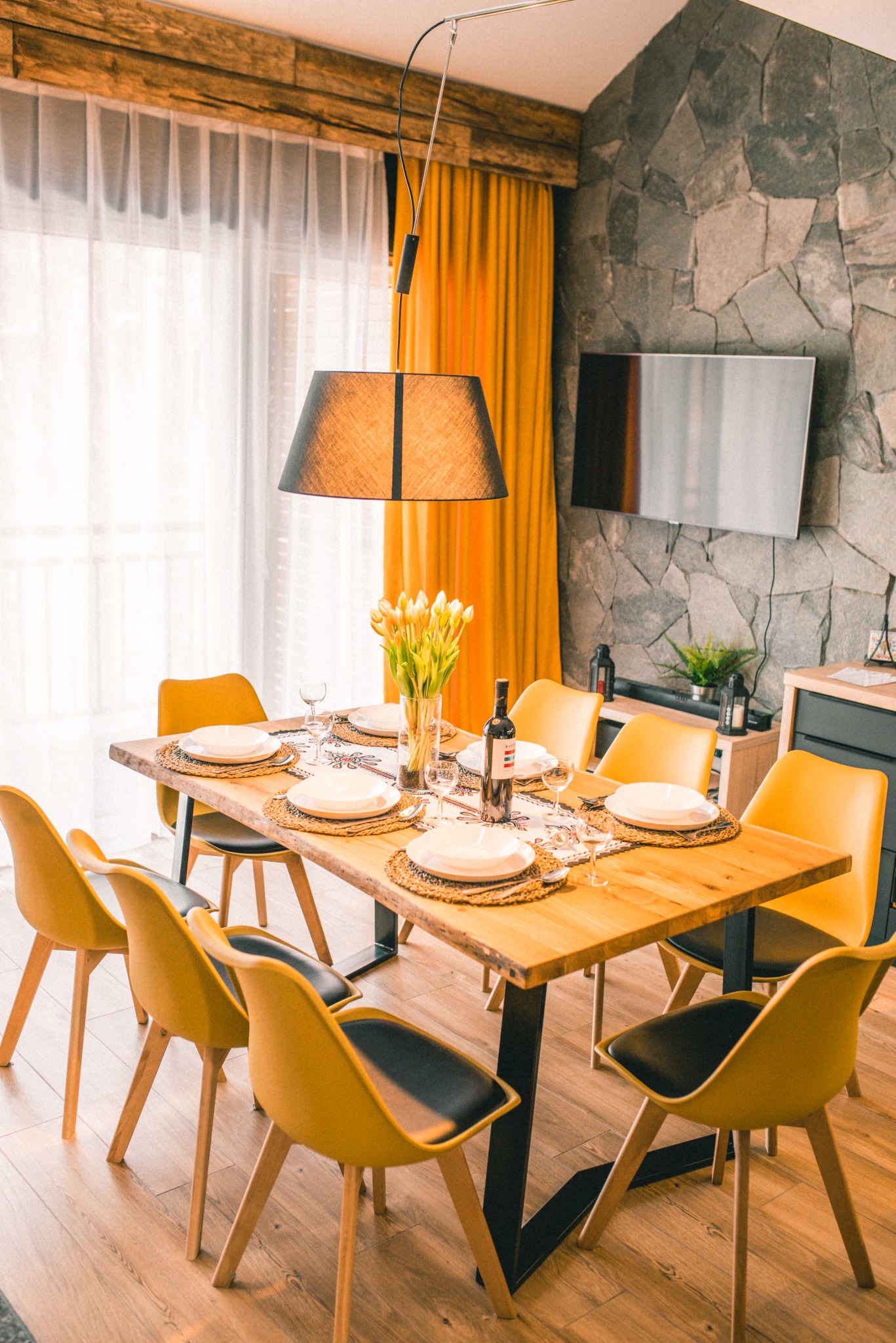 Jadalnia z drewnianym stołem, żółtymi krzesłami i kamienną ścianą. Stół nakryty do posiłku, w wazonie tulipany. Żółte zasłony w oknie. Nowoczesny wystrój.