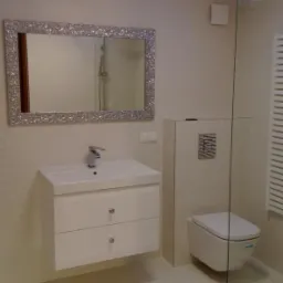 Nowoczesna łazienka w jasnych barwach z lustrem w ozdobnej ramie, podświetleniem LED sufitu i kabiną prysznicową ze szkła.