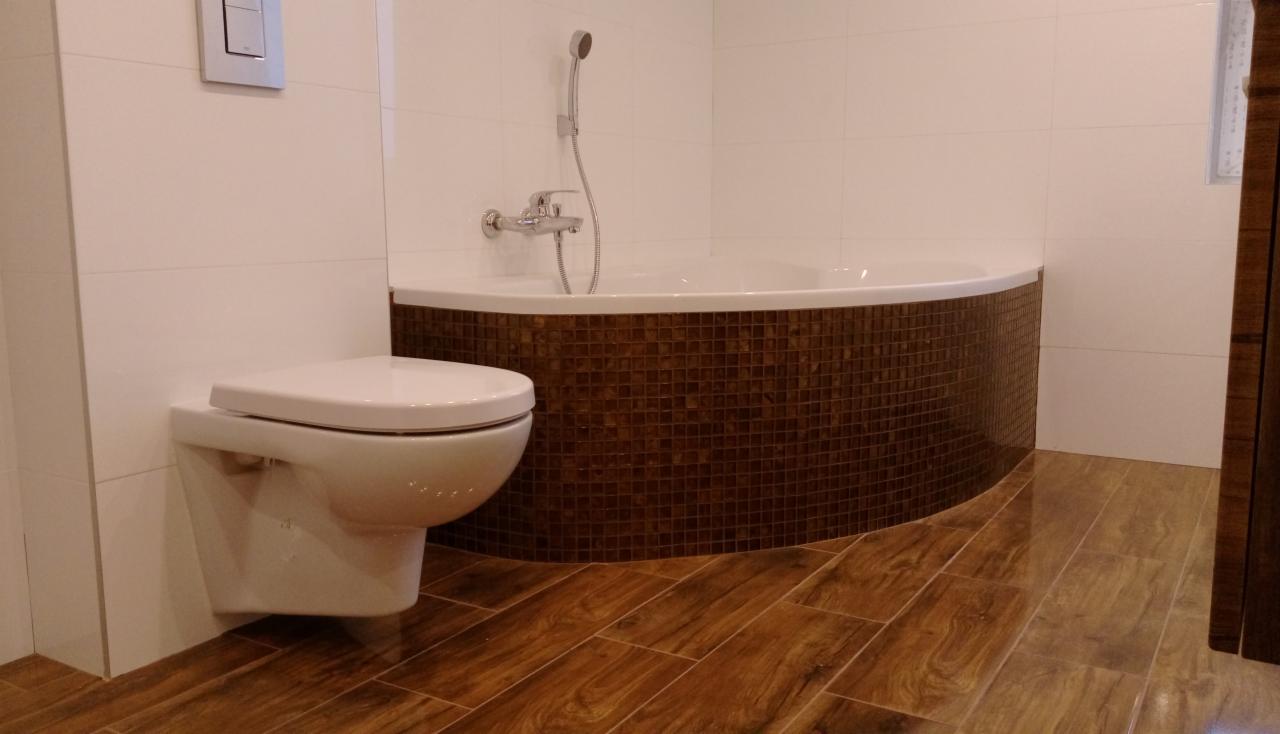 Nowoczesna łazienka z brązową mozaiką na obudowie wanny, białą armaturą i podłogą imitującą drewno, widok na wiszącą miskę WC i baterię prysznicową.