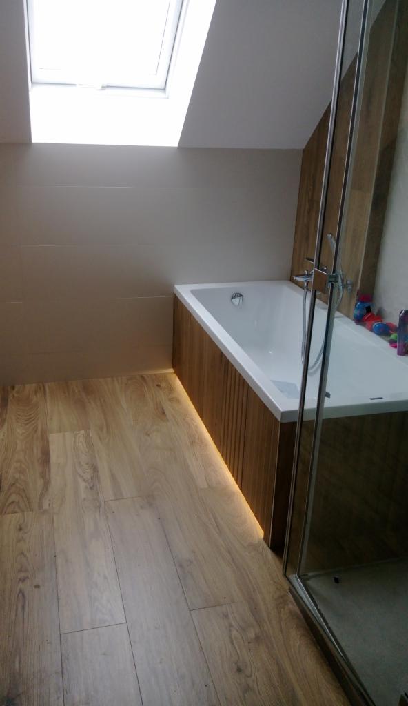 Nowoczesna łazienka na poddaszu z oknem dachowym, drewnianą obudową wanny z podświetleniem LED, drewnopodobną podłogą i częściowo przeszkloną kabiną prysznicową.