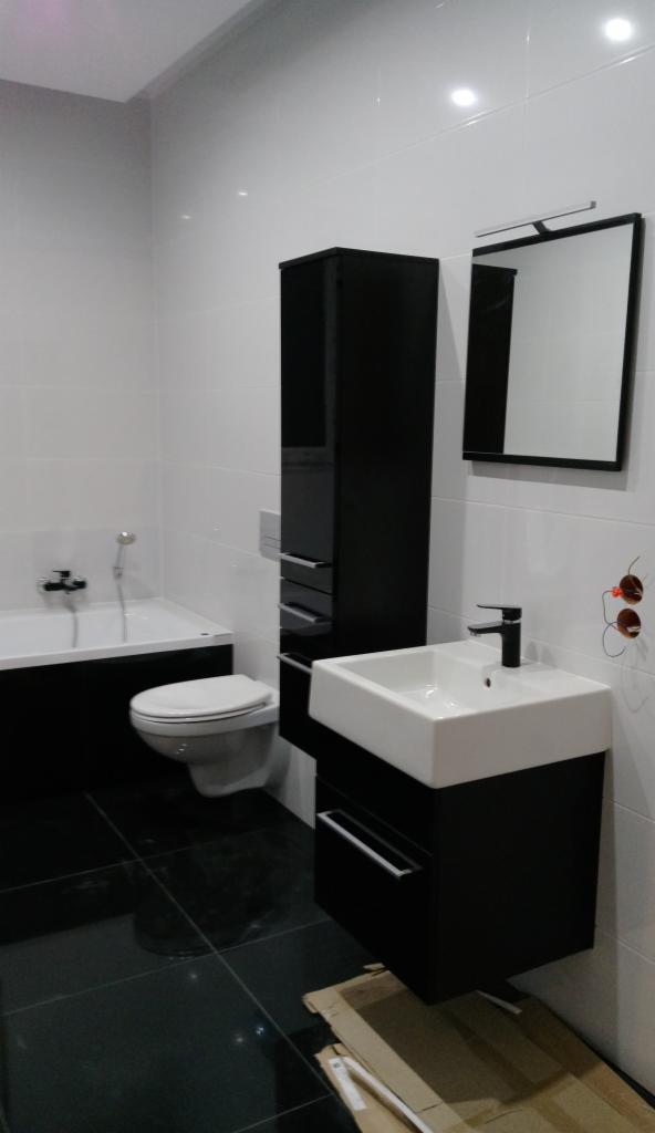 Nowoczesna łazienka w czarno-białej kolorystyce: białe płytki ścienne, czarna podłoga, wanna z czarną obudową, wisząca szafka z umywalką, wysoka szafka łazienkowa oraz lustro z oświetleniem.