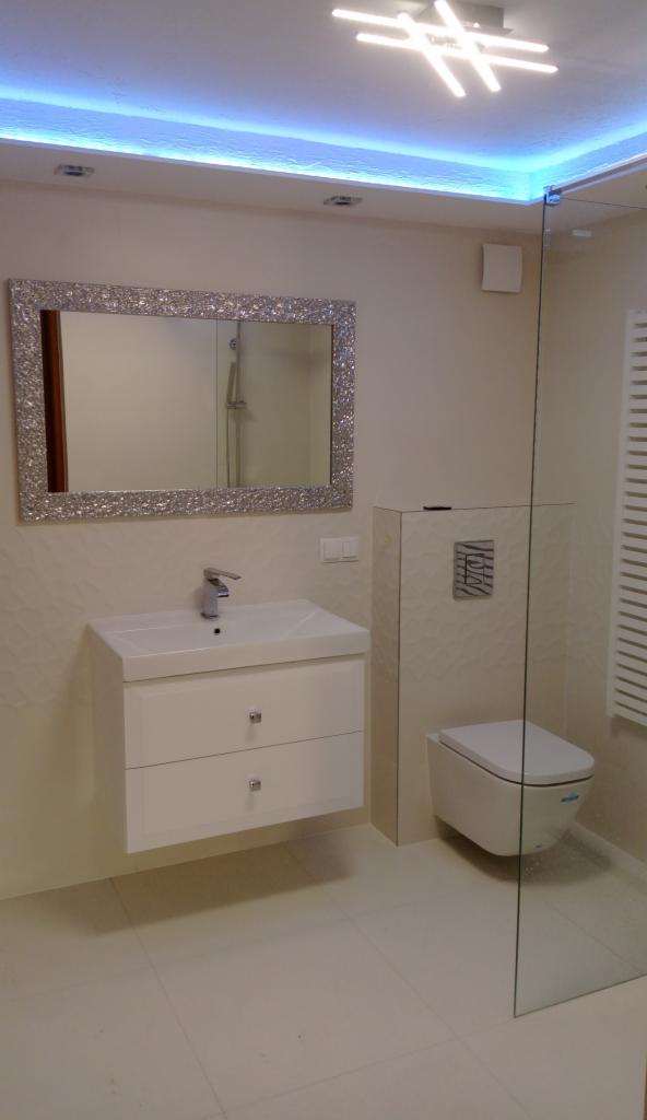 Nowoczesna łazienka w jasnych barwach z lustrem w ozdobnej ramie, podświetleniem LED sufitu i kabiną prysznicową ze szkła.